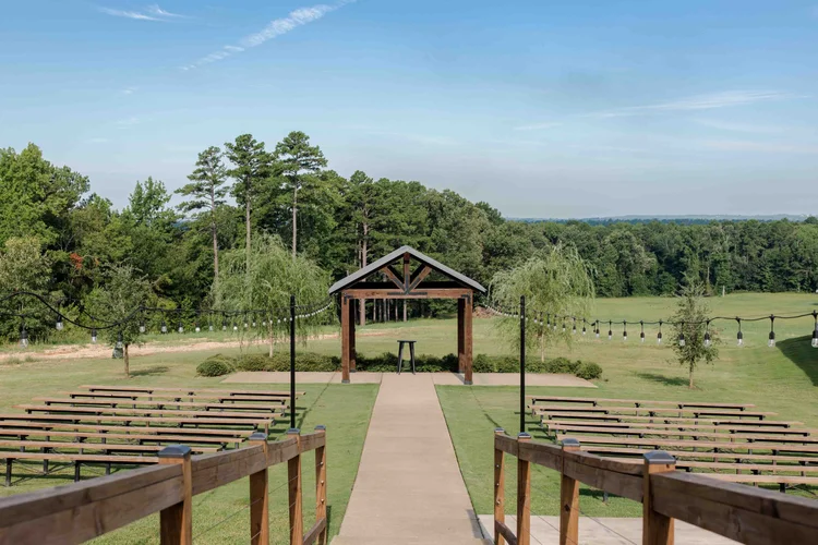 Outdoor wedding ceremony setup with wooden chairs arranged on either side of a central aisle leading to a small wooden chapel, surrounded by string lights and greenery, with a backdrop of trees and a blue sky.