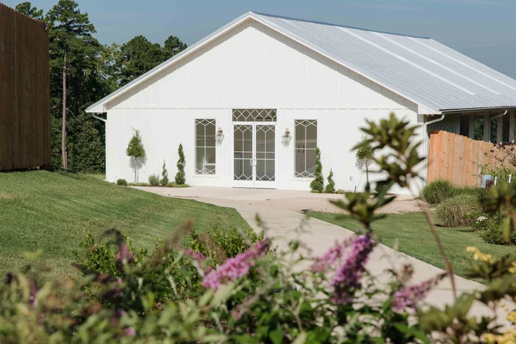 A white house with a sloped metal roof, double doors, and three windows, surrounded by a garden and a paved pathway.